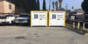 Two portable storage containers labeled "Box Go Storage" placed in a driveway in Carthay Square, with utility trucks and palm trees in the background.