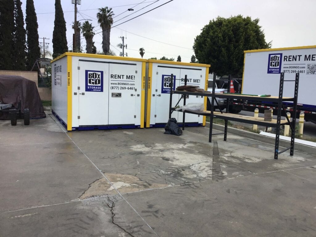 Three Box-n-Go storage containers on a paved lot in Koreatown, Los Angeles, with metal shelving in the foreground