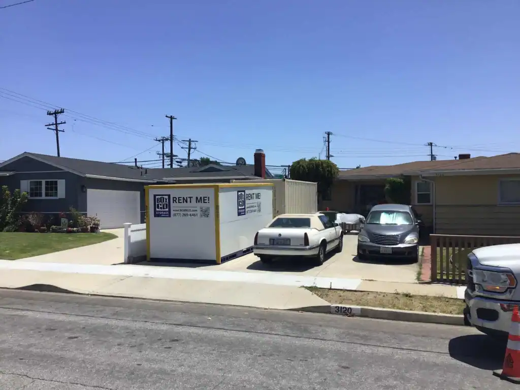 Playa Vista Movers storage container parked in residential driveway during moving day, with cars and homes in the background under clear blue skies.