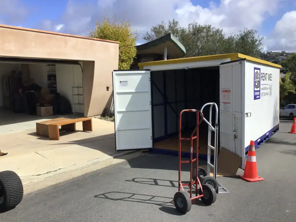Homeowners loading boxes into a portable storage unit beside an open garage, illustrating convenient alternatives for those who want to rent a garage for extra storage or moving needs.