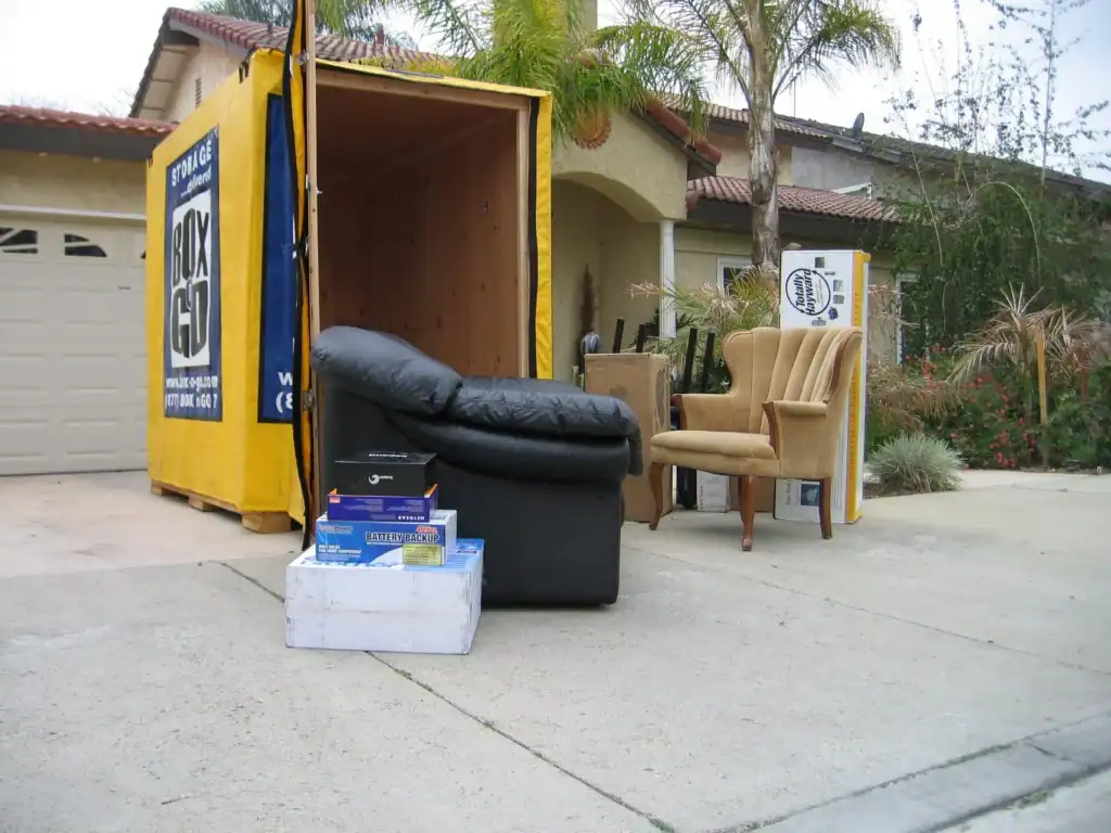 A BOX-GO STORAGE container, vibrant in yellow and blue, is parked in a residential driveway, its door open to reveal a wooden interior.