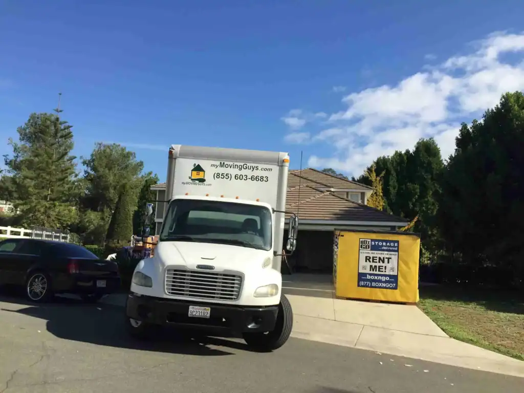 Moving truck parked in front of a suburban home with a yellow Box-n-Go portable container in the driveway, highlighting convenient and affordable Self Storage Unit in Calabasas CA for residential moving and storage solutions.