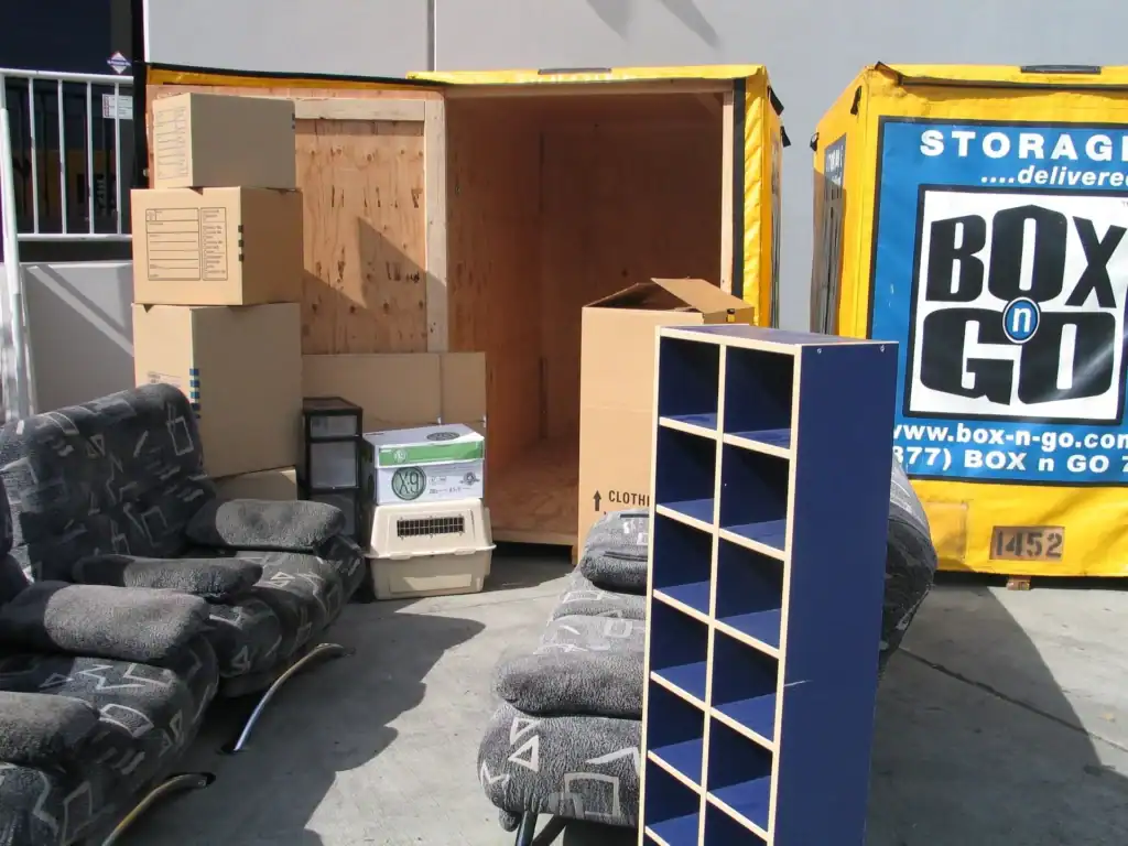 Packing fragile items for storage with sturdy moving boxes and protective furniture placement beside portable storage containers, showing organized preparation for safe transport and storage of delicate belongings.