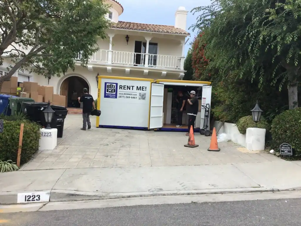 Two individuals are actively involved in moving operations, either loading or unloading items from a "BOX-GO STORAGE" container, which prominently displays "RENT ME!" and contact information.