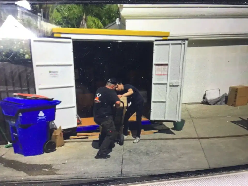 Two movers carefully packing fragile items for storage into a portable container, ensuring safe handling and secure loading at a residential driveway.
