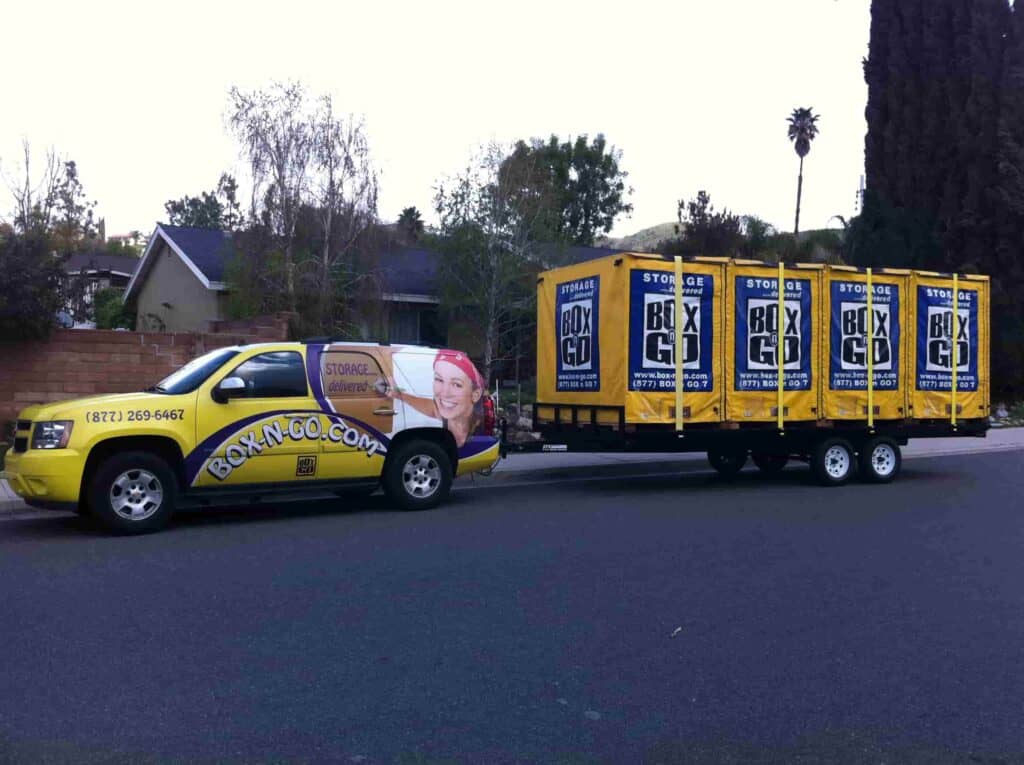 A branded Box-n-Go truck hauls multiple yellow portable storage containers through a residential neighborhood, highlighting convenient and flexible self-storage-valley-village services for local homeowners.