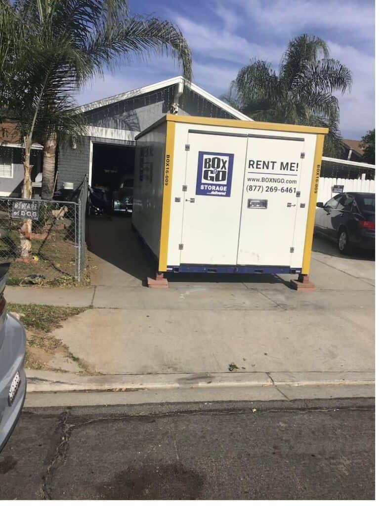 White Box-n-Go storage container on residential driveway with garage of vintage cars behind