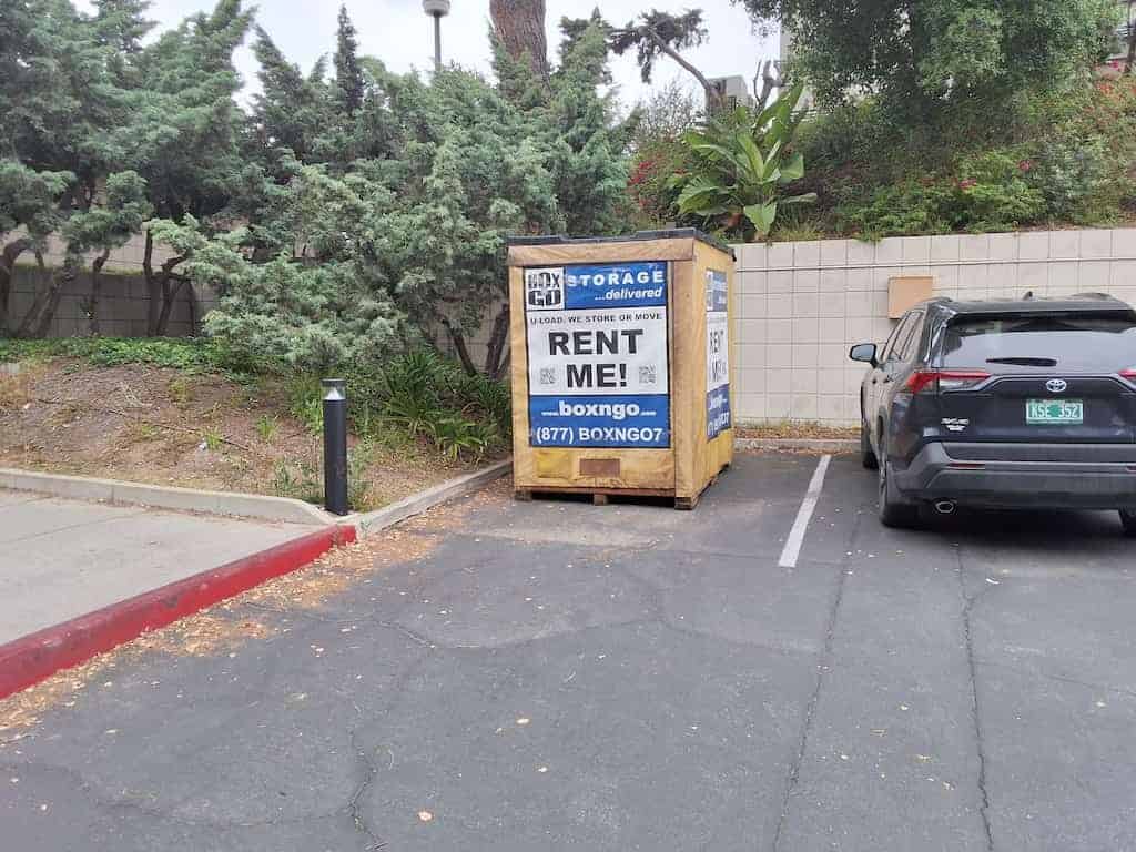 A plywood storage vault standing in a parking bay beside a landscaped slope of shrubs, dropped there for student storage near CalArts.