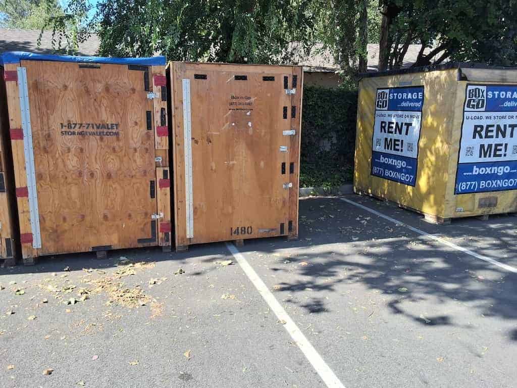 Two plywood storage vaults and two yellow Box-n-Go containers parked together in a shaded lot, the equipment delivered for student storage near The Masters University.