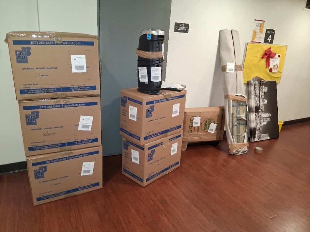 Labeled cartons stacked on a wood floor with a rolled rug, framed pictures and a tall cylindrical appliance beside them, cleared out for Vanguard University dorm storage.