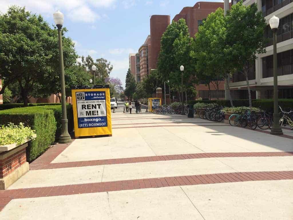 Two Box-n-Go containers on a paved campus walkway lined with clipped hedges during move-out week, one at the near end and one further down, in use for The Master's University student storage.