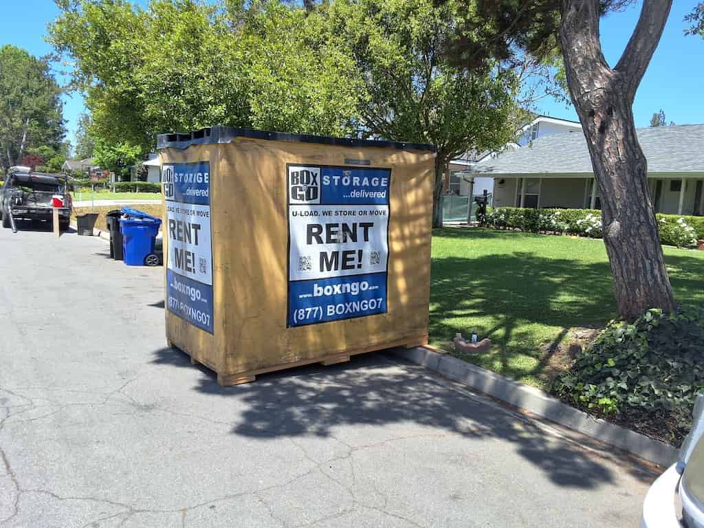 A covered Box-n-Go container standing on a residential driveway beside a lawn and a tree, delivered to an off-campus house for CalArts summer storage.