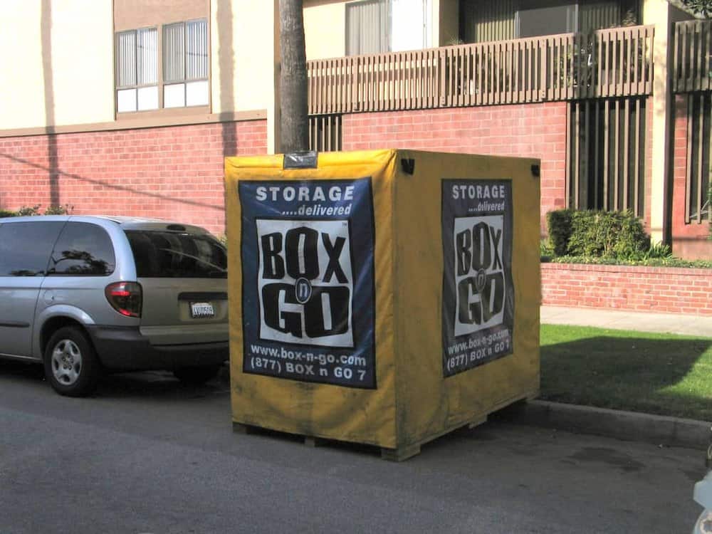 A yellow Box-n-Go container standing at the curb outside a brick apartment building with a parked minivan beside it, delivered for Vanguard University summer storage.