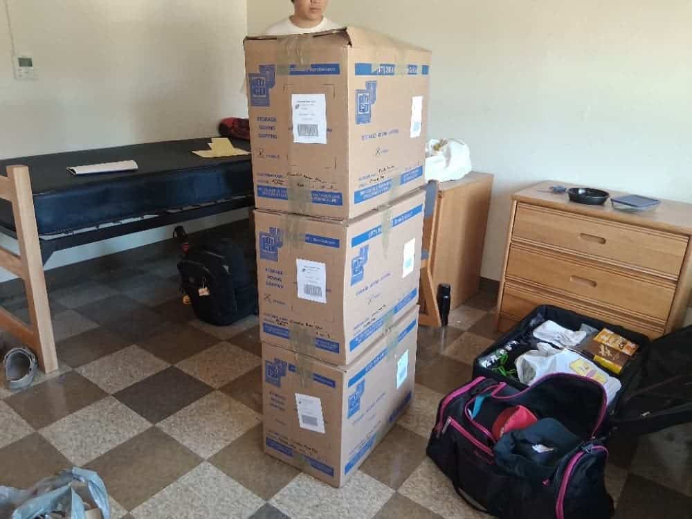 A stack of three labeled cartons in a residence room beside a stripped bed frame, a dresser and two open bags, ready for CalArts dorm storage.