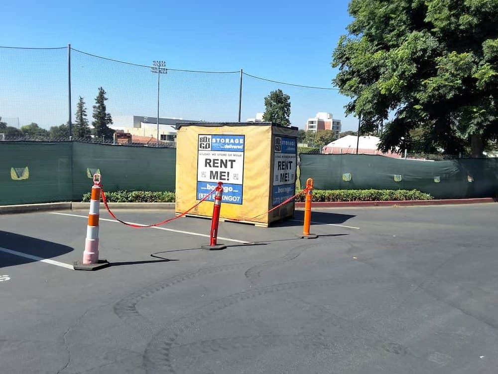 A yellow Box-n-Go container coned off with traffic cones and tape in a parking lot beside green tennis court fencing, held for Orange Coast College summer storage.