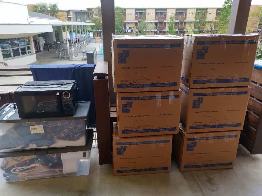 Labeled cartons, a black microwave, clear bins and a glass-front cabinet staged on an apartment walkway above a courtyard, waiting for storage units near CalArts.