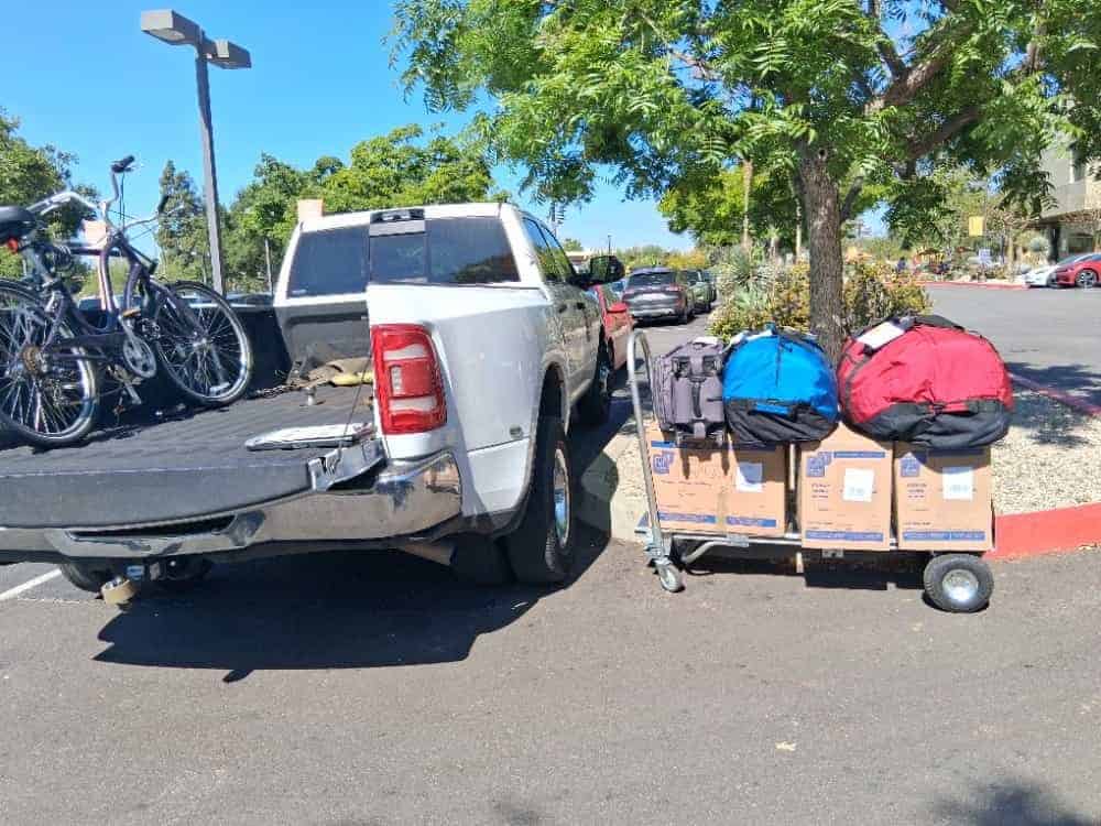 Bicycles in a pickup bed and a flat cart of labeled cartons and duffel bags waiting in a parking lot on TMU move out storage day.