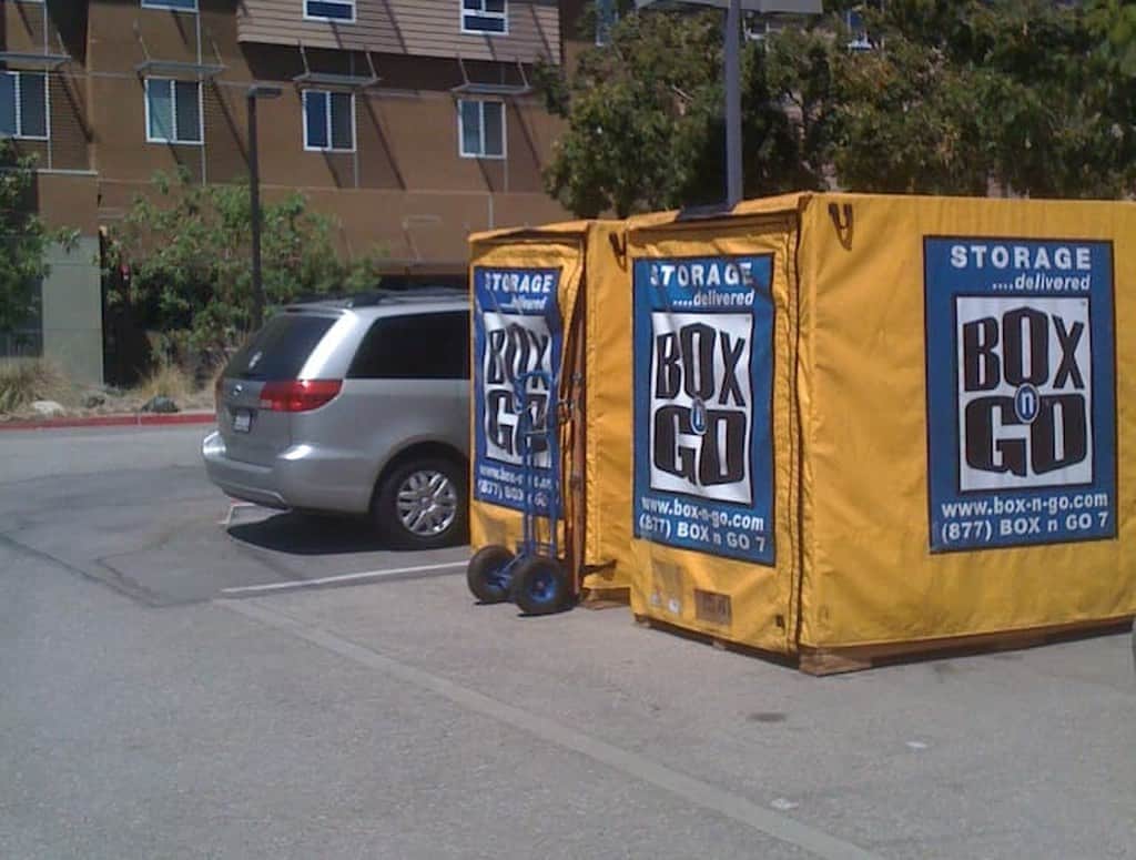 Two yellow Box-n-Go move out storage containers standing in a parking bay outside a campus residence hall, a hand truck between them.